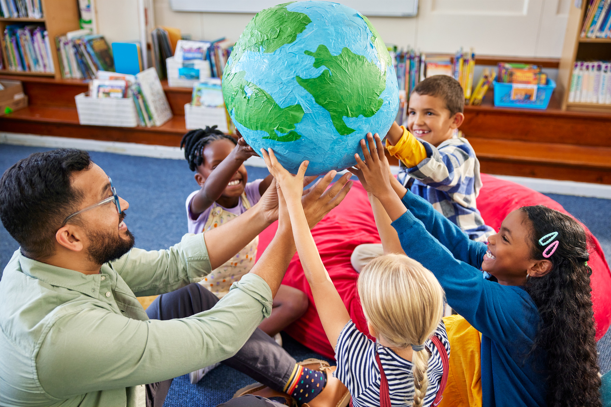 Group of multiethnic children with male teacher holding model of earth in classroom. High angle view of geography teacher with schoolgirls and schoolboys holding a big handmade earth model. Hands of children holding a planet model while studying about the world, earth day and environment friendly concept.